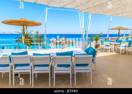 Spiaggia di Cala Nova, isola di Ibiza - 20 Maggio 2017: Bianco tavolo e sedie con cuscini blu sulla terrazza ristorante sulla spiaggia di Cala Nova con belle vie del mare Foto Stock