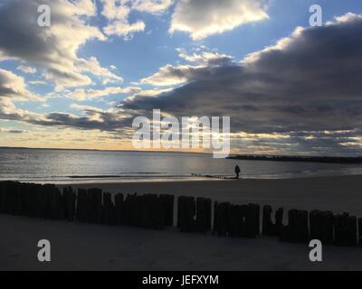 La persona cammina da sola lungo la spiaggia sabbiosa al tramonto. Le nuvole drammatiche si sweep attraverso il cielo. Resti di un molo di legno, una fila di pali di legno intemperie Foto Stock