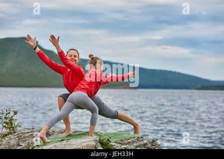 Giovane madre e figlia piccola pratica yoga di bilanciamento pongono sulla roccia vicino al fiume. Foto Stock