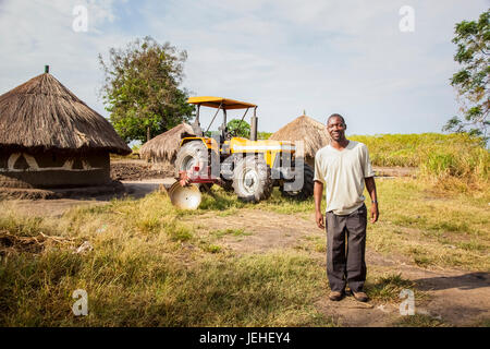 Un uomo si erge in posa con un trattore e capanna in background; Uganda Foto Stock
