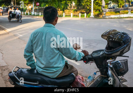 Un uomo si siede sulla sua moto a lato della strada come un ciclo rickshaw approcci; Krong Siem Reap, Siem Reap Provincia, Cambogia Foto Stock