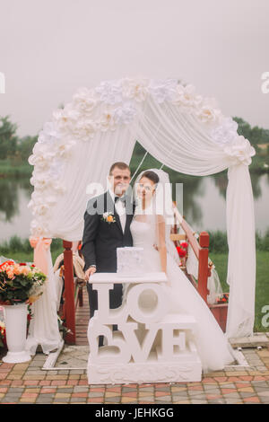 Il Sorridente sposi sono in piedi dietro il segno di nozze e arch. Foto Stock