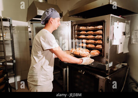 Bello baker in uniforme tenendo il vassoio pieno di pane appena sfornato a livello di fabbricazione Foto Stock