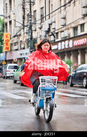 Ragazza cinese in rosso antipioggia su un e-bike. Shanghai è un umido clima subtropicale, l'estate è molto caldo e umido. Foto Stock