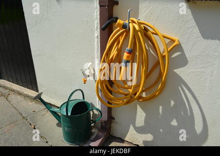 Giardino giallo tubo hanging off rivestito in plastica gancio di metallo al di fuori del dipinto di garage con metallo verde annaffiatoio sul patio a lastroni sotto acqua corrente fredda Foto Stock