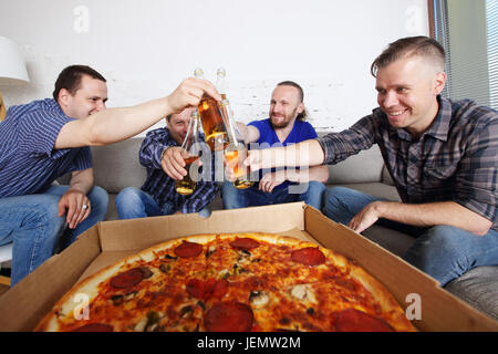 Amici maschi seduti a casa sul lettino, il Clink birra, andando a mangiare la pizza, celebrando qualcosa Foto Stock