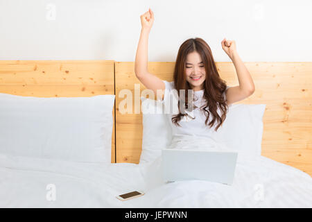 Felice giovane bella donna asiatica svegliarsi nel suo letto, eccitato il tifo al suo schermo di computer Foto Stock