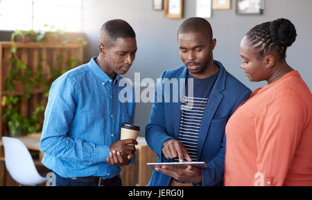 Tre focalizzata giovane africana office i colleghi parlare insieme su una tavoletta digitale mentre in piedi in un grande ufficio moderno Foto Stock