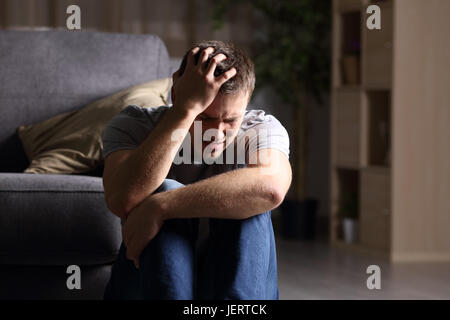 Triste uomo piange seduto sul pavimento del salotto di una casa interna con uno sfondo scuro Foto Stock