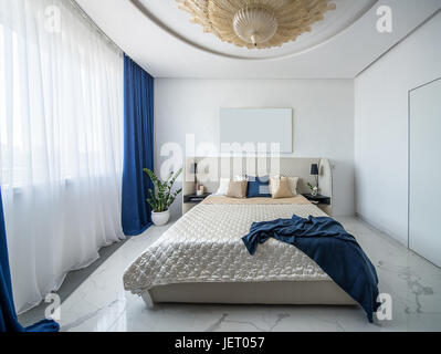 Camera moderna con una grande fantasia lampadario dorato sul soffitto. Vi è un letto di coloro beige con multicolore di cuscini e un blu plaid, mensole con candela Foto Stock