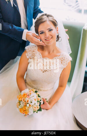 Incantevole sposa si siede nella poltrona azienda bouquet nuziali e amare lo sposo si erge nei pressi di Foto Stock