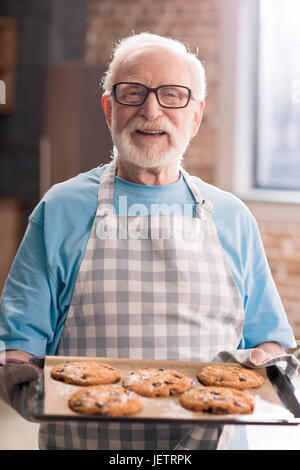 Senior uomo grembiule nella vaschetta di contenimento di deliziosi biscotti, cottura in concetto di cucina Foto Stock