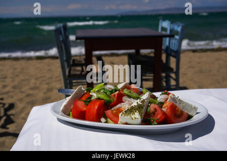 Insalata Xoriatiki servita su un tavolo da spiaggia greca con sfocate taverna sulla spiaggia tavolo in legno e sedie blu sullo sfondo. Foto Stock