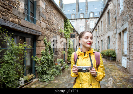 Giovane donna in giallo impermeabile a piedi con zaino e macchina fotografica a Dinan villaggio alla regione della Bretagna in Francia Foto Stock