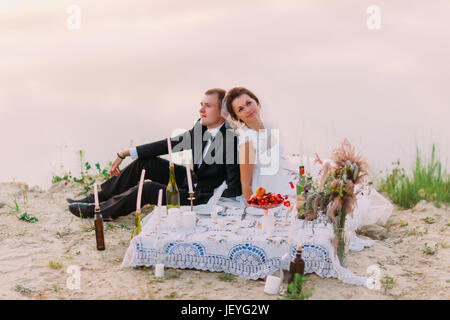 Il romantico ritratto orizzontale degli sposi sirring back-to-back pur avendo il wedding picnic sulla costa il wedding picnic sulla costa Foto Stock