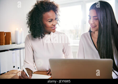 Due multirazziale giovane femmina business partner seduto a un computer portatile di sorridere quando essi pianificare la loro strategia di business insieme come un team Foto Stock