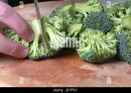 Close up di una mano di un uomo di trinciatura broccoli lavati con un coltello in acciaio su un tagliere di legno. Alcune piccole goccioline di acqua visibile. Foto Stock