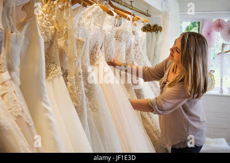 Un assistente di vendita in un abito da sposa shop, guardando attraverso il abiti appesi nelle rotaie. Foto Stock