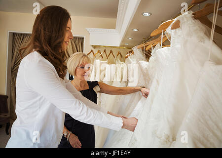 Due donne, una giovane sposa di essere e una matura assistente vendite guardando attraverso le rotaie di abiti da sposa. Foto Stock