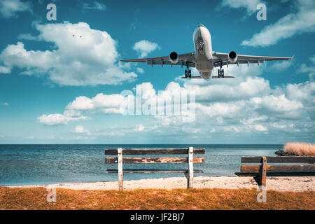 Splendido aeroplano e panca di legno al mare. Paesaggio con bianco aereo passeggeri vola nel cielo blu con nuvole sopra il mare in estate. Pass Foto Stock