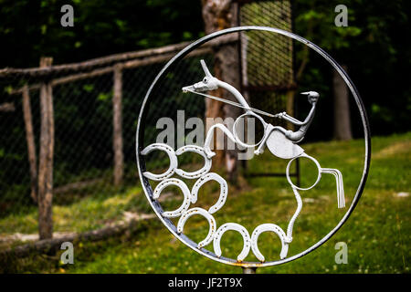 Segno bianco con un cavaliere e ferri di cavallo in piedi su erba con la recinzione e foresta in background. Foto Stock