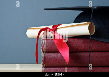 Close up di un mortarboard e graduazione scorrere sulla sommità di una pila di vecchi libri usurati, posto su di un legno chiaro Tavolo con uno sfondo grigio. Foto Stock