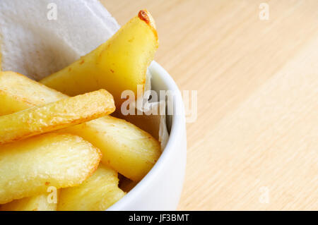 Close up (macro) di una ciotola di chips (patatine fritte) adagiata su carta assorbente in una ciotola bianco. Tavolo in legno superficie fornisce spazio copia a destra Foto Stock