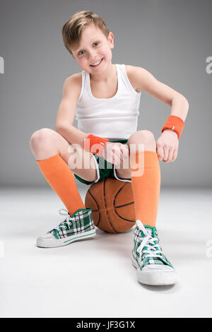 Ragazzo sorridente nella seduta di abbigliamento sportivo sul basket ball isolato su grigio Foto Stock