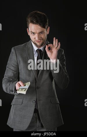 Giovane imprenditore tenendo dollar banconote e mostra ok segno isolato su nero Foto Stock