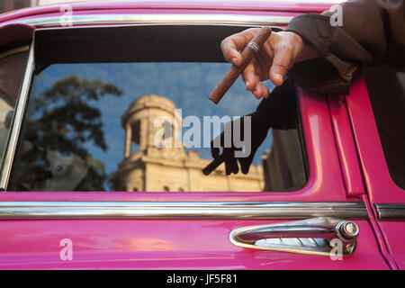 Un uomo detiene un sigaro come architettura cubana è riflessa nella finestra di un colorato classic American car. Foto Stock