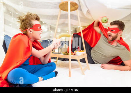 Entusiasta il padre ed il figlio in costumi di supereroi divertendosi con frutti in coperta fort Foto Stock