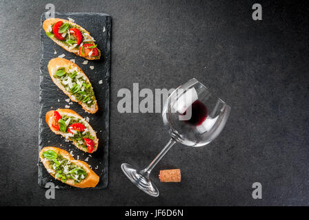 Il cibo italiano. Spuntini. Antipasto. Bruschetta con pesto, parmigiano, pomodori e basilico fresco. Sulla tavola nera, sulla scheda di ardesia. Con bicchiere di Foto Stock
