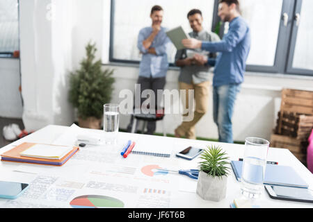 Vista ravvicinata di grafici aziendali e delle forniture di ufficio sulla scrivania e di imprenditori in piedi dietro, business concetto del lavoro di squadra Foto Stock