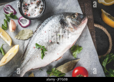 Crudo fresco pesce organico Dorado, orata con gli ingredienti per la cottura di limone, i pomodori, la cipolla, il sale, il pepe e la foglia di alloro, verdi, olio. Sul vecchio cutti in legno Foto Stock