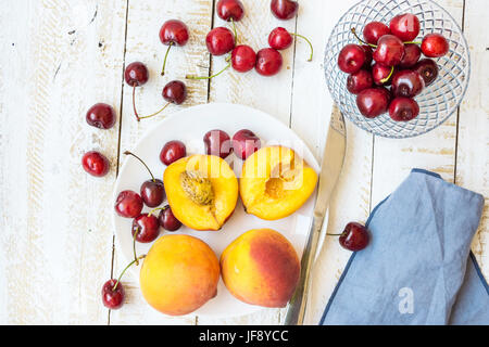 Colorate mature le pesche tagliate a metà e interi di ciliegie dolci sulla piastra bianca, ciotola di cristallo, coltello, plank tavolo in legno blu, igienico, di stile e disposizione, la piana Foto Stock