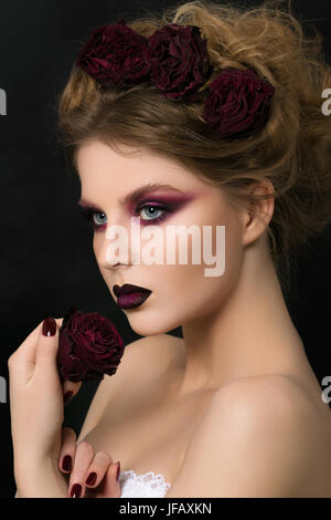 Close-up ritratto di giovane donna con viola scuro partito make-up holding rosso scuro rose appassite Foto Stock