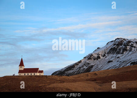 Semplice chiesa con un tetto rosso e steeple a VIK, Islanda attraverso un marrone campo di erba con una coperta di neve montagna in background e cielo blu e nuvole Foto Stock