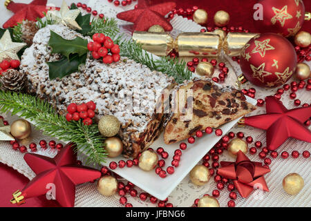 Natale tradizionale stollen di cioccolato la torta su un piatto con una slice, rosso e oro decorazioni ninnolo, lamina di cioccolatini incartati, santo e fir. Foto Stock