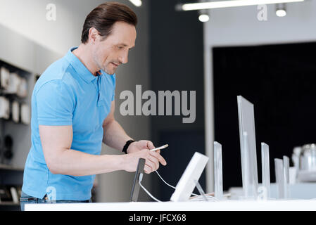 Guardando le caratteristiche principali. Interessati adulto uomo indossare una polo blu in piedi vicino a un negozio display mentre lo shopping per l'ultimo telefono cellulare Foto Stock