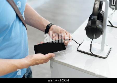 Permettetemi di provare questo fuori. Su scala di colpo di un cliente maschio cercando di plug mockup cuffie nel suo smartphone mentre si sta in piedi in un display e shopping f Foto Stock