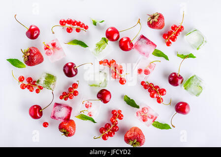 Vista dall'alto di cubetti di ghiaccio con frutti di bosco freschi tra non congelati ciliegia, fragola e menta leafs sullo sfondo bianco. Messa a fuoco selettiva Foto Stock