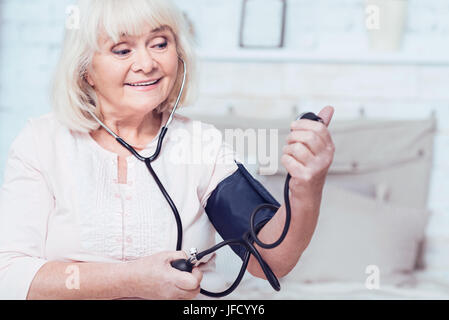 Controllare la mia salute. Sorridente età affascinante donna seduta sul letto di casa e misurare la pressione del sangue mentre esprimono gioia Foto Stock