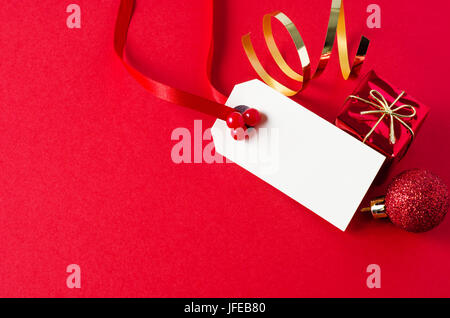 Un bianco Natale regalo tag, decorata con un nastro rosso e artificiale di bacche di agrifoglio, su una superficie opaca con uno sfondo rosso brillante piccola scatola regalo, glittery baubl Foto Stock