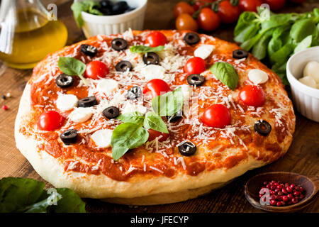 Pizza con pomodori, mozzarella, olive nere e basilico. Una deliziosa pizza italiana su legno pizza board. Vista ingrandita, il fuoco selettivo Foto Stock