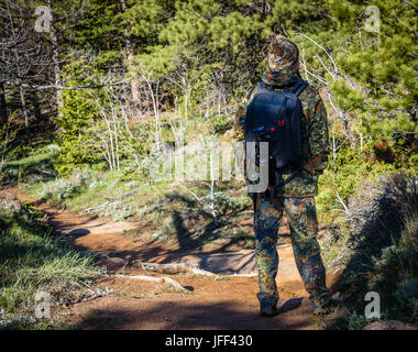 Escursionista uomo fotografo in camuffamento vestito con uno zaino e treppiede in piedi su una montagna sentiero forestale e guardare la fauna selvatica, vista posteriore Foto Stock