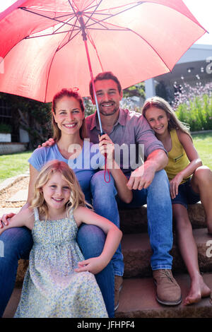 La famiglia felice seduti in giardino sotto un ombrello rosso Foto Stock