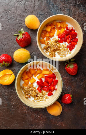 Lo yogurt naturale con pezzi di Albicocche Fragole, granola e pinoli in due ciotole su broun scuro dello sfondo. Vista superiore Foto Stock