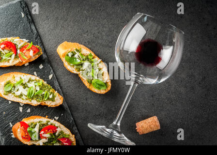 Il cibo italiano. Spuntini. Antipasto. Bruschetta con pesto, parmigiano, pomodori e basilico fresco. Sulla tavola nera, sulla scheda di ardesia. Con bicchiere di Foto Stock