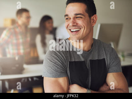 Sorridente fiducioso giovane uomo con le braccia incrociate e permanente che guarda lontano dall'ufficio. Foto Stock