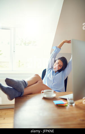Stretching giovane donna con espressione stanchi e piedi sulla scrivania in ufficio a casa. Tazza di caffè e computer in primo piano. Foto Stock